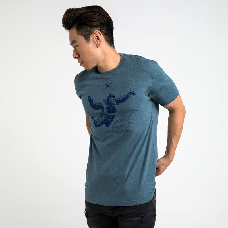 May include: A teal blue t-shirt with a graphic of a gorilla wearing a parachute and being pulled by a drone over a city skyline.
