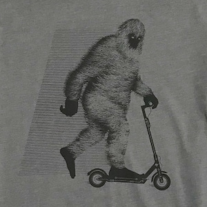 May include: Gray t-shirt featuring a graphic of a Bigfoot riding a black scooter. The Bigfoot is depicted in shades of gray with a furry texture, and the scooter has a modern design. A striped design is in the background.