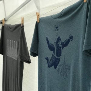 May include: A teal blue t-shirt with a graphic print of a gorilla wearing a parachute and flying over a city skyline. The gorilla is holding a remote control in one hand and a drone is flying above it.