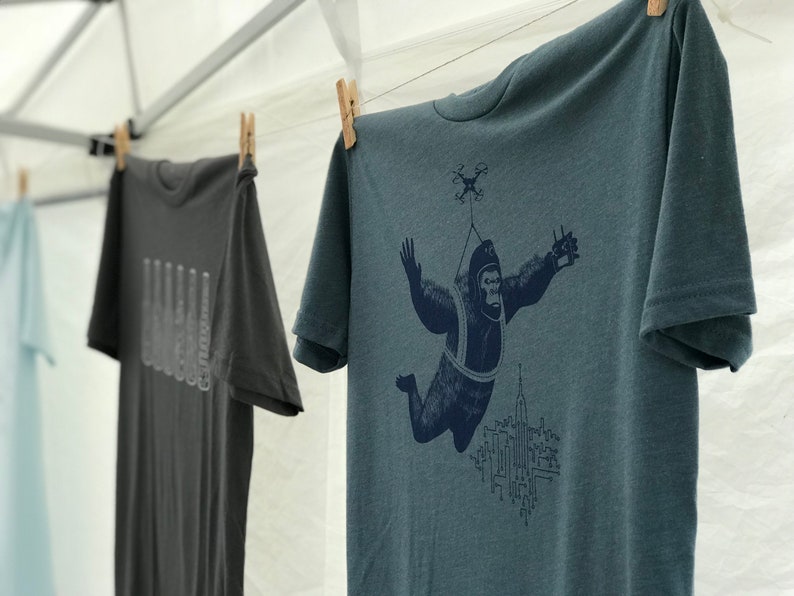 May include: A teal blue t-shirt with a graphic print of a gorilla wearing a parachute and flying over a city skyline. The gorilla is holding a remote control in one hand and a drone is flying above it.
