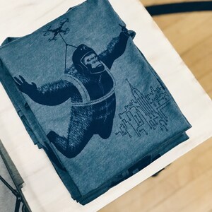 May include: A stack of blue t-shirts with a graphic print of a gorilla wearing a helmet and a harness, holding onto a drone. The gorilla is flying over a cityscape with circuit board designs.