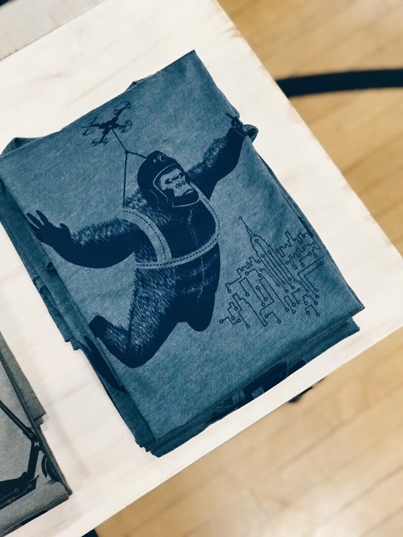 May include: A stack of blue t-shirts with a graphic print of a gorilla wearing a helmet and a harness, holding onto a drone. The gorilla is flying over a cityscape with circuit board designs.