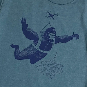 May include: A blue graphic t-shirt featuring a design of a gorilla wearing a helmet and harness, flying with a drone above a city skyline. The gorilla is holding a remote control in one hand.