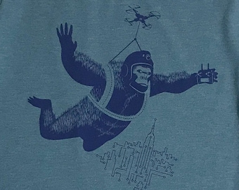 May include: A blue graphic tee shirt with a design of a gorilla wearing a helmet and a harness, flying with a drone above a city skyline. The gorilla is holding a remote control in one hand.