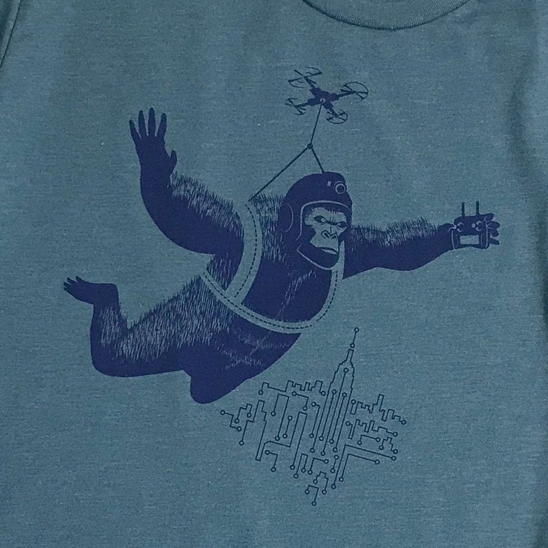 May include: A blue graphic tee shirt with a design of a gorilla wearing a helmet and a harness, flying with a drone above a city skyline. The gorilla is holding a remote control in one hand.