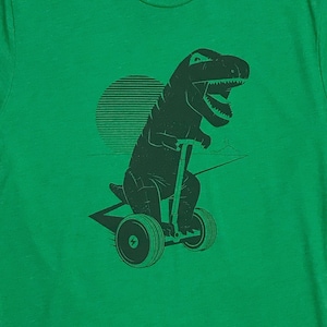May include: A green t-shirt with a black silhouette of a dinosaur riding a hoverboard. The dinosaur has a large mouth and is facing forward. The hoverboard has two wheels and a handle.