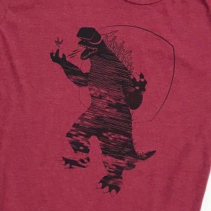 Może przedstawiać: Czerwony T-shirt z czarną grafiką potwora w wirtualnej rzeczywistości. Potwór trzyma mały przedmiot w ręce i ma groźny wyraz twarzy.