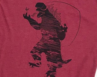 May include: A red t-shirt with a black graphic of a monster wearing a virtual reality headset. The monster is holding a small object in its hand and has a menacing expression.