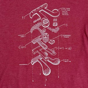 Może przedstawiać: Bordowa koszulka z białym schematycznym wzorem symbolu Pi. Projekt zawiera liczby, r&oacute;wnania i tekst "Pi Day: March 14th". Unikalna koszulka graficzna dla entuzjast&oacute;w matematyki.