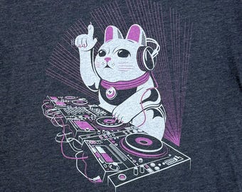 May include: Dark gray t-shirt featuring a white cat DJ with headphones, a purple scarf, and a raised finger. The cat is positioned in front of a DJ mixer with purple accents and a radiating purple design.