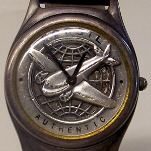 May include: Silver-toned wristwatch with a black leather band. The watch face features a detailed airplane design over a globe, with the word "AUTHENTIC" around the edge. The watch has a classic, vintage aesthetic.