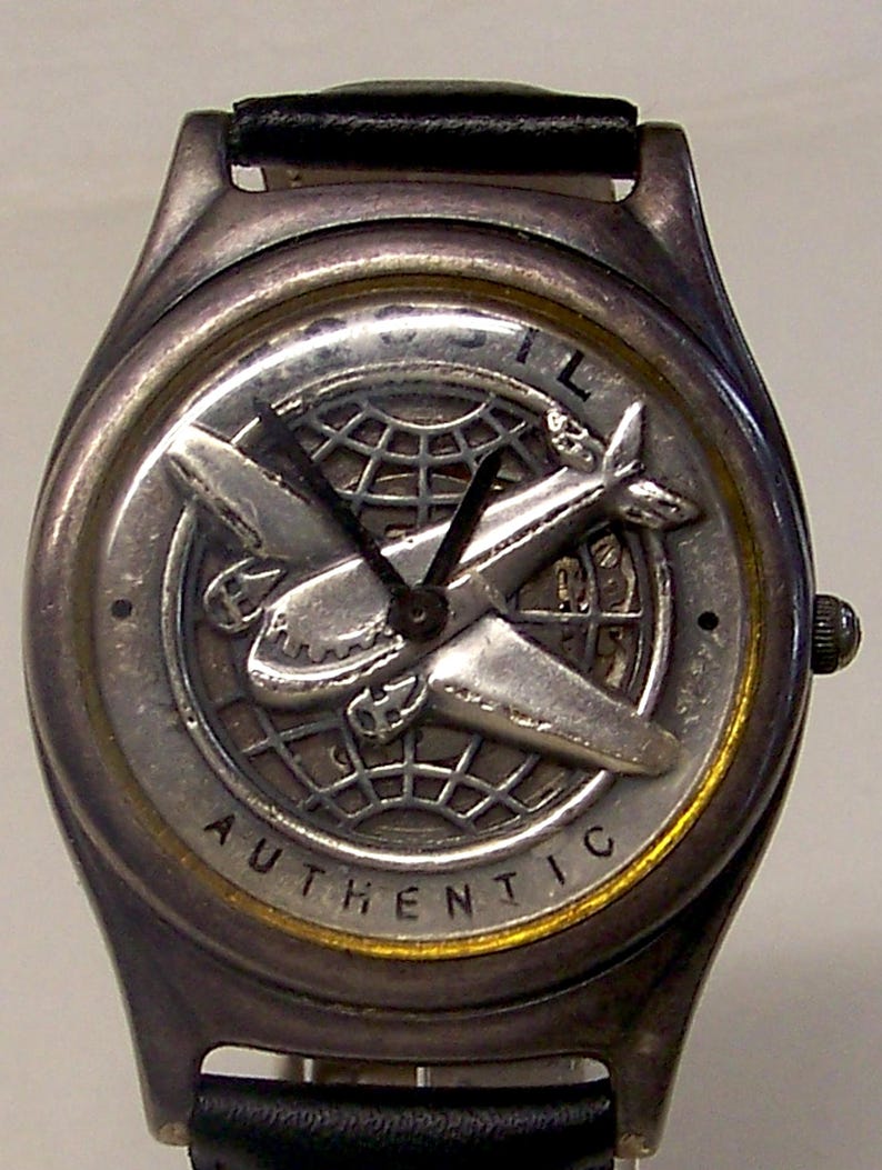 May include: Silver-toned wristwatch with a black leather band. The watch face features a detailed airplane design over a globe, with the word "AUTHENTIC" around the edge. The watch has a classic, vintage aesthetic.