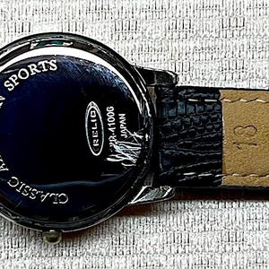 Puede incluir: Correa de reloj de cuero negro con hebilla plateada. La esfera del reloj es azul oscuro con las palabras "AMERICAN SPORTS CLASSIC" y un logotipo. La correa est&aacute; marcada con "WATER PROOF" y "Genuine Leather".