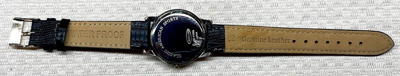 Puede incluir: Correa de reloj de cuero negro con hebilla plateada. La esfera del reloj es azul oscuro con las palabras "AMERICAN SPORTS CLASSIC" y un logotipo. La correa est&aacute; marcada con "WATER PROOF" y "Genuine Leather".