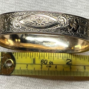 May include: A gold-toned hinged bangle bracelet featuring an intricate engraved floral and leaf pattern with a central marquise-shaped motif. A yellow measuring tape is positioned underneath, showing the bracelet width is approximately 2.25 inches.