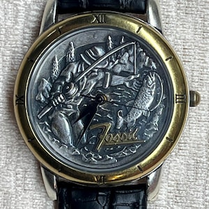 May include: A Fossil brand wristwatch with a silver and gold-tone case and a black leather band. The watch face features a detailed relief scene of a fisherman, trees, and a fish. The watch has Roman numeral hour markers.