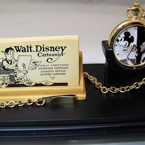 May include: A gold pocket watch with a black and white image of Mickey Mouse and Walt Disney on the face. The watch is on a black stand with a gold chain. A wooden card holder with a black and white image of Walt Disney drawing is also on the stand.