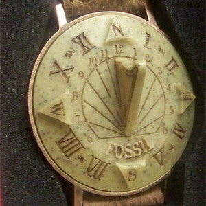 May include: A Fossil brand wristwatch with a cream-colored face and a brown leather band. The face features a compass design with Roman numerals and a sundial in the center.