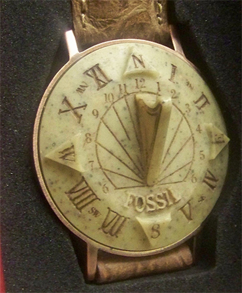 May include: A Fossil brand wristwatch with a cream-colored face and a brown leather band. The face features a compass design with Roman numerals and a sundial in the center.