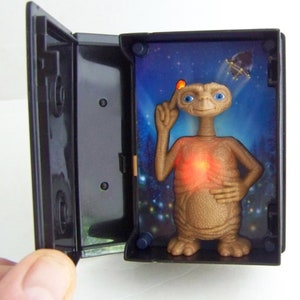 May include: A black cassette tape case with a light brown alien figure inside. The alien is holding its finger to its head and has a red light on its chest. The background is a dark blue sky with stars and a flying saucer.