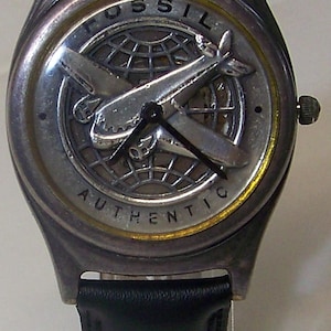 May include: A Fossil watch with a silver-tone case and a black leather band. The watch face features a silver airplane design over a globe, with the word "FOSSIL" at the top and "AUTHENTIC" at the bottom.