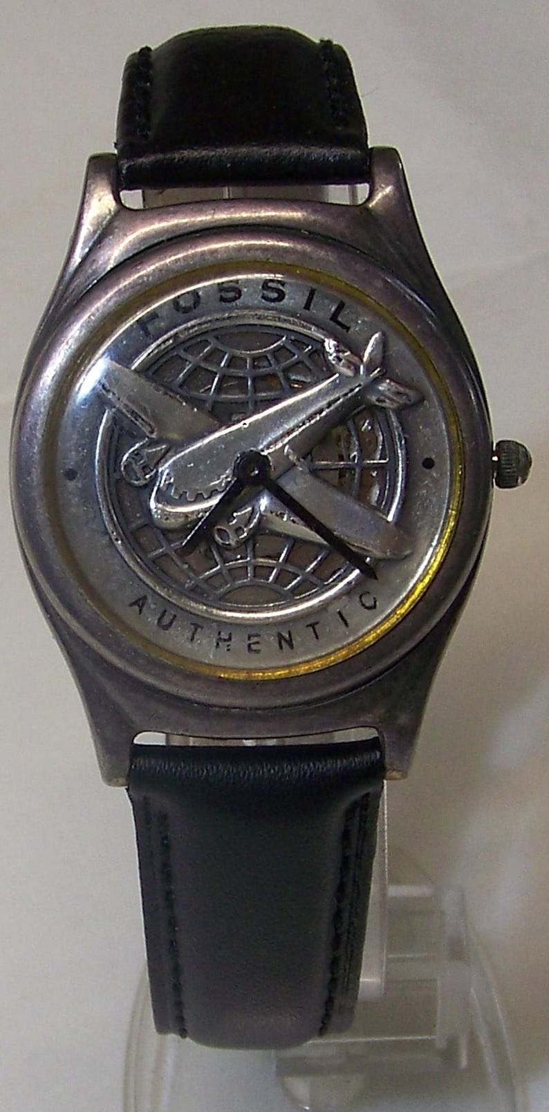 May include: A Fossil watch with a silver-tone case and a black leather band. The watch face features a silver airplane design over a globe, with the word "FOSSIL" at the top and "AUTHENTIC" at the bottom.