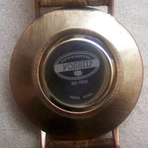 May include: The back of a round, silver-toned watch with a black face. The watch face has the word "FOSSIL" printed in white letters on a silver oval. The watch face also has the number "SO-7620" printed in white letters.