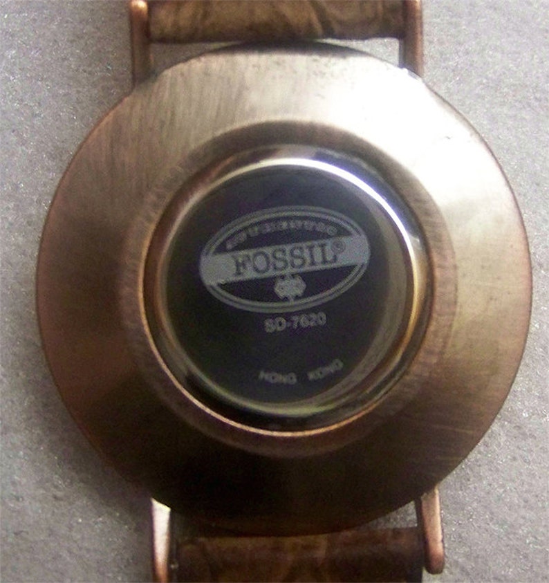 May include: The back of a round, silver-toned watch with a black face. The watch face has the word "FOSSIL" printed in white letters on a silver oval. The watch face also has the number "SO-7620" printed in white letters.