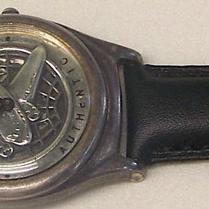 May include: A wristwatch with a black leather band and a silver-toned watch face. The watch face features a detailed design. The band is secured with a silver buckle.