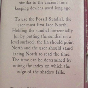 May include: A close-up of a white card with instructions on how to use a Fossil Sundial. The card has the text "You have just purchased the Fossil Sundial." and "To use the Fossil Sundial, the user must first face North." The card also includes the phone number 1-800-TIC-TOCI.