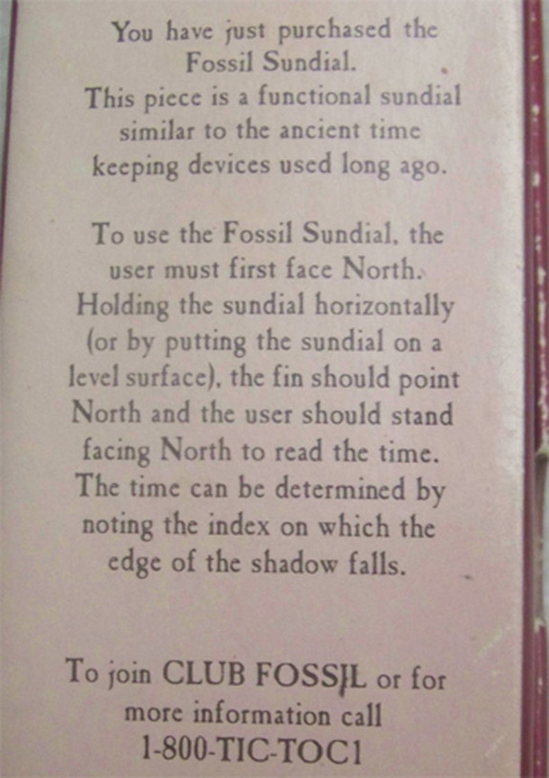 May include: A close-up of a white card with instructions on how to use a Fossil Sundial. The card has the text "You have just purchased the Fossil Sundial." and "To use the Fossil Sundial, the user must first face North." The card also includes the phone number 1-800-TIC-TOCI.