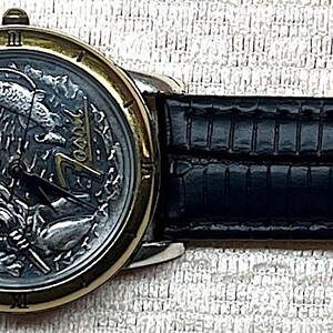 Puede incluir: Un reloj de pulsera con una correa de cuero negro y una esfera plateada y dorada. La esfera presenta un dise&ntilde;o detallado. La correa mide aproximadamente 2 cm de ancho.