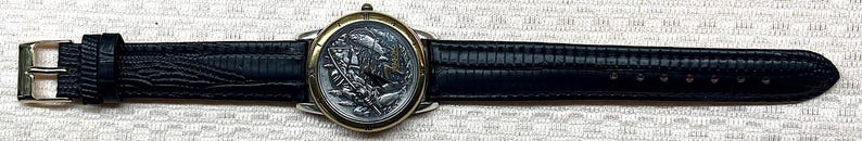 Puede incluir: Un reloj de pulsera con una correa de cuero negro y una esfera plateada y dorada. La esfera presenta un dise&ntilde;o detallado. La correa mide aproximadamente 2 cm de ancho.