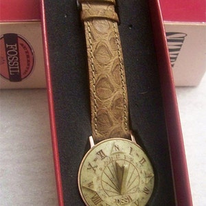 May include: A brown leather wristwatch with a sundial face. The watch face is white with black Roman numeral markings and a gold-colored metal frame. The watch is in a red box with the word "Fossil" printed on it.