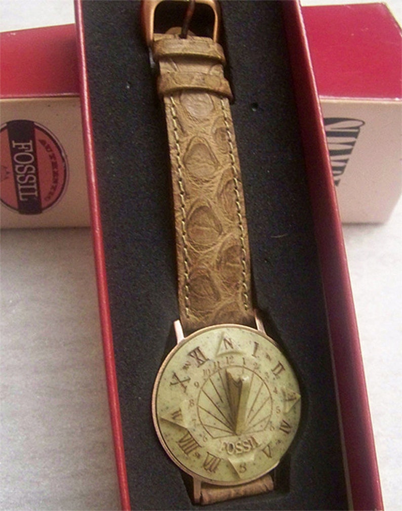 May include: A brown leather wristwatch with a sundial face. The watch face is white with black Roman numeral markings and a gold-colored metal frame. The watch is in a red box with the word "Fossil" printed on it.