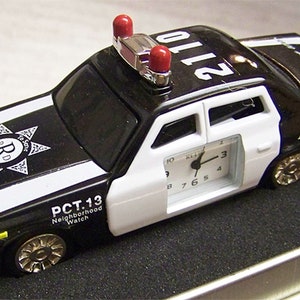 May include: Black and white toy police car with a clock on the side. The car has a red and silver light on top and says "Dial 911 Emergency" on the back. The car also has a star with "R.D. Reserve Deputy" written on it and "PCT. 13 Neighborhood Watch" on the side.