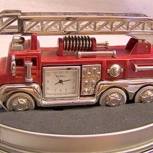 May include: A red and silver fire truck clock with a ladder on top. The clock face is square with black numbers and hands. The clock is set inside a metal tin.