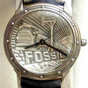 May include: A silver Fossil wristwatch with a black leather band. The watch face features a detailed engraving of a train and the word "Fossil".