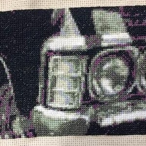 Op de afbeelding: Een kruissteek borduurwerk op beige Aida-stof met de voorkant van een oldtimer. Het ontwerp heeft een zwarte achtergrond met een gedetailleerde chromen bumper, rechthoekige en ronde koplampen en een grille in grijstinten.