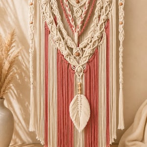 May include: A macrame wall hanging featuring cream and terracotta colored cotton cords suspended from a wooden dowel. The design includes intricate knotted patterns, vertical fringe sections, wooden beads, and a central leaf-shaped element.