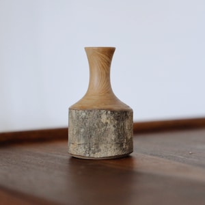 May include: A small wooden vase with a natural bark finish. The vase has a narrow neck and a wider base, and it is made from a single piece of wood.