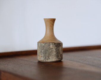 May include: A small wooden vase with a natural bark finish. The vase has a narrow neck and a wider base, and it is made from a single piece of wood.