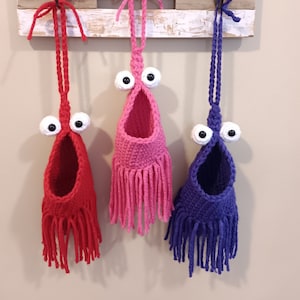 May include: Three handmade crochet wall pockets in red, pink, and purple, each with large googly eyes and fringe. They hang from a wooden sign that reads "Home Sweet Home."