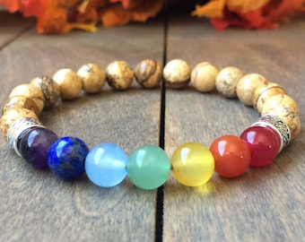 Puede incluir: Un brazalete de chakra arcoíris con siete cuentas de colores y un brazalete de piedra marrón moteada. El brazalete está sobre una superficie de madera.