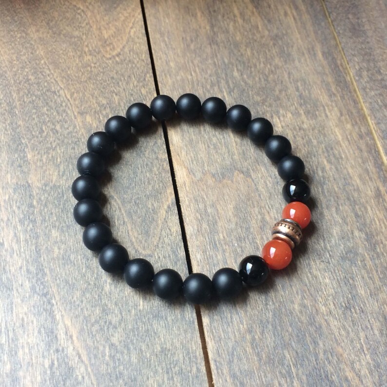 A black matte beaded bracelet with two orange carnelian beads and a copper spacer bead.