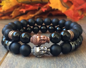 Puede incluir: Dos pulseras de cuentas negras. Una pulsera presenta una cabeza de Buda de cobre, y la otra tiene una cabeza de Buda plateada. Las pulseras están hechas de cuentas negras mate y brillantes, con detalles de cobre y plata.