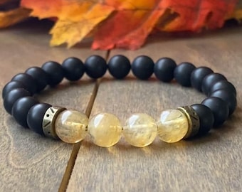 Puede incluir: Un brazalete de cuentas negras y doradas con tres cuentas de color amarillo dorado y dos separadores dorados. El brazalete está sobre una superficie de madera.