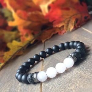 May include: A black and white beaded bracelet with silver accents. The bracelet features black matte beads, white beads, and lava rock beads.