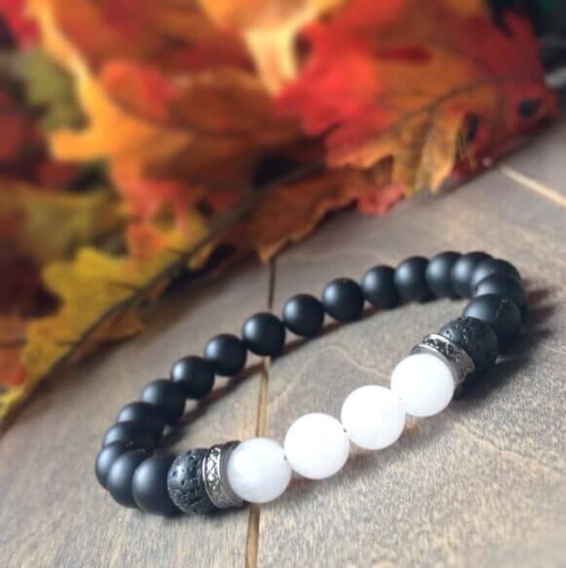 May include: A black and white beaded bracelet with silver accents. The bracelet features black matte beads, white beads, and lava rock beads.