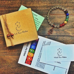 May include: A clear crystal bracelet with a hamsa charm and seven chakra colored beads. The bracelet is packaged in a brown box with the text "Unlock Your Chakra" on it. The box is sitting on a wooden surface.